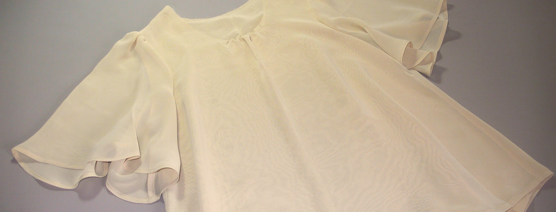 A flared blouse made of chiffon in a shade of ochre laid out on a gray table. 