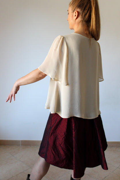 A woman photographed from the back wearing a short-sleeved chiffon blouse with a flared shape in shades of ochre and a voluminous midi Bordeaux skirt.
