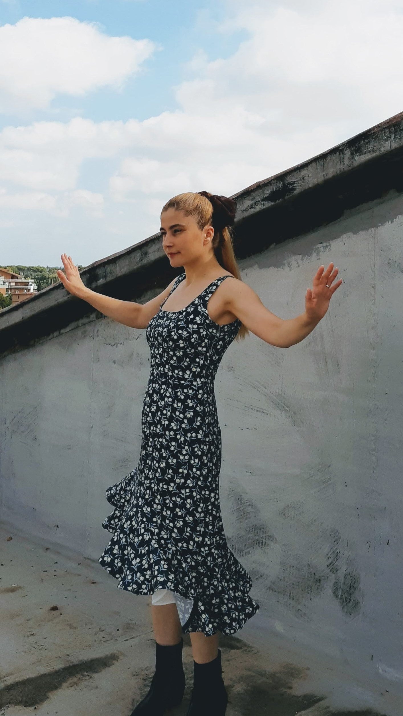 A woman with a ponytail posing in a flouncy sleeveless patterned dress.