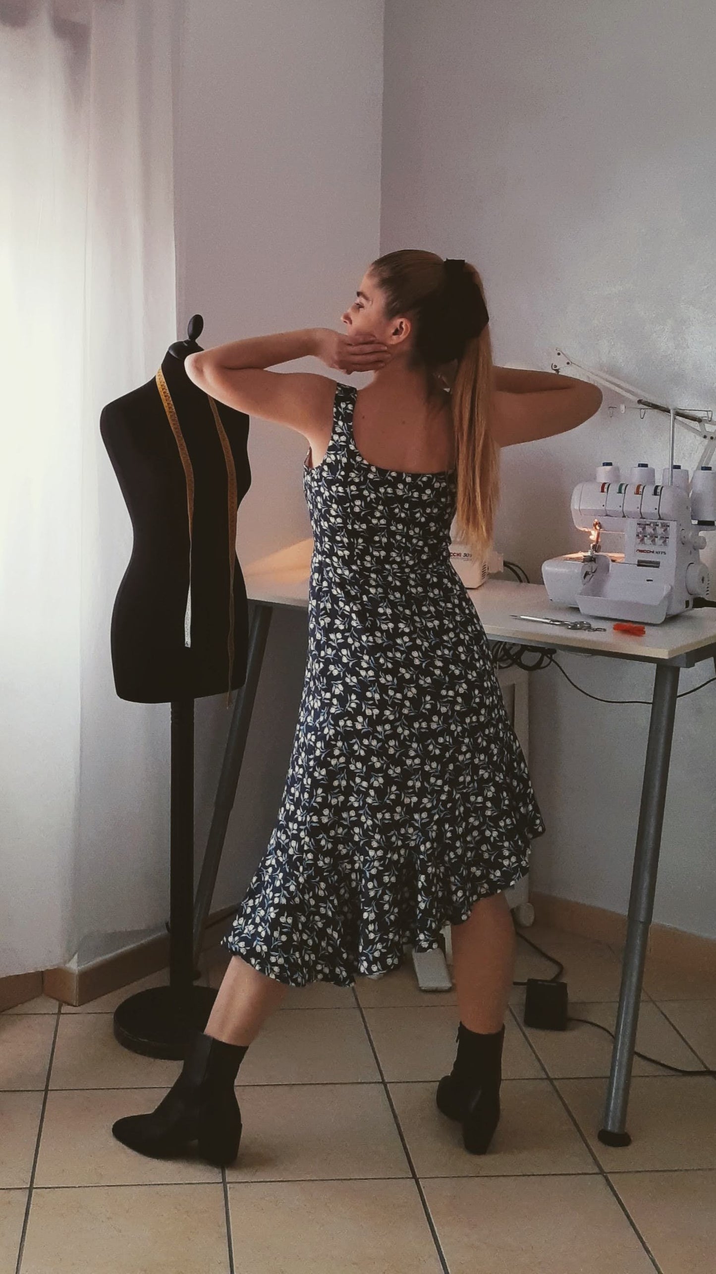 A woman with a ponytail is posing in a asymmetric sleeveless dress and black boots. The view is from the back.