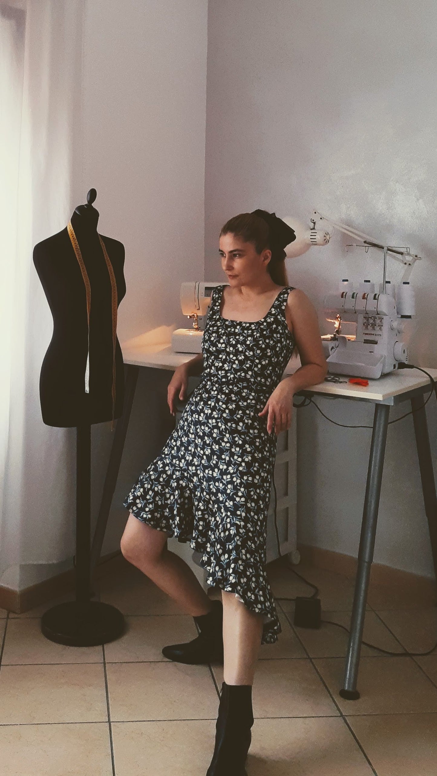 A woman in a patterned dress poses beside a tailoring mannequin amidst sewing machines on a table in the background.
