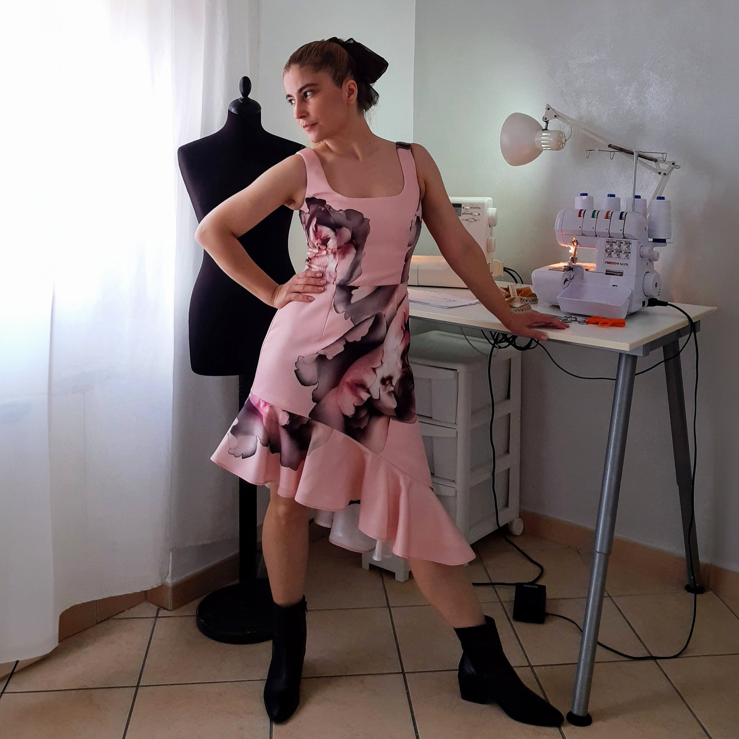 A woman in a pink asymmetric dress poses beside a tailor mannequin amidst sewing machines on a table in the background.