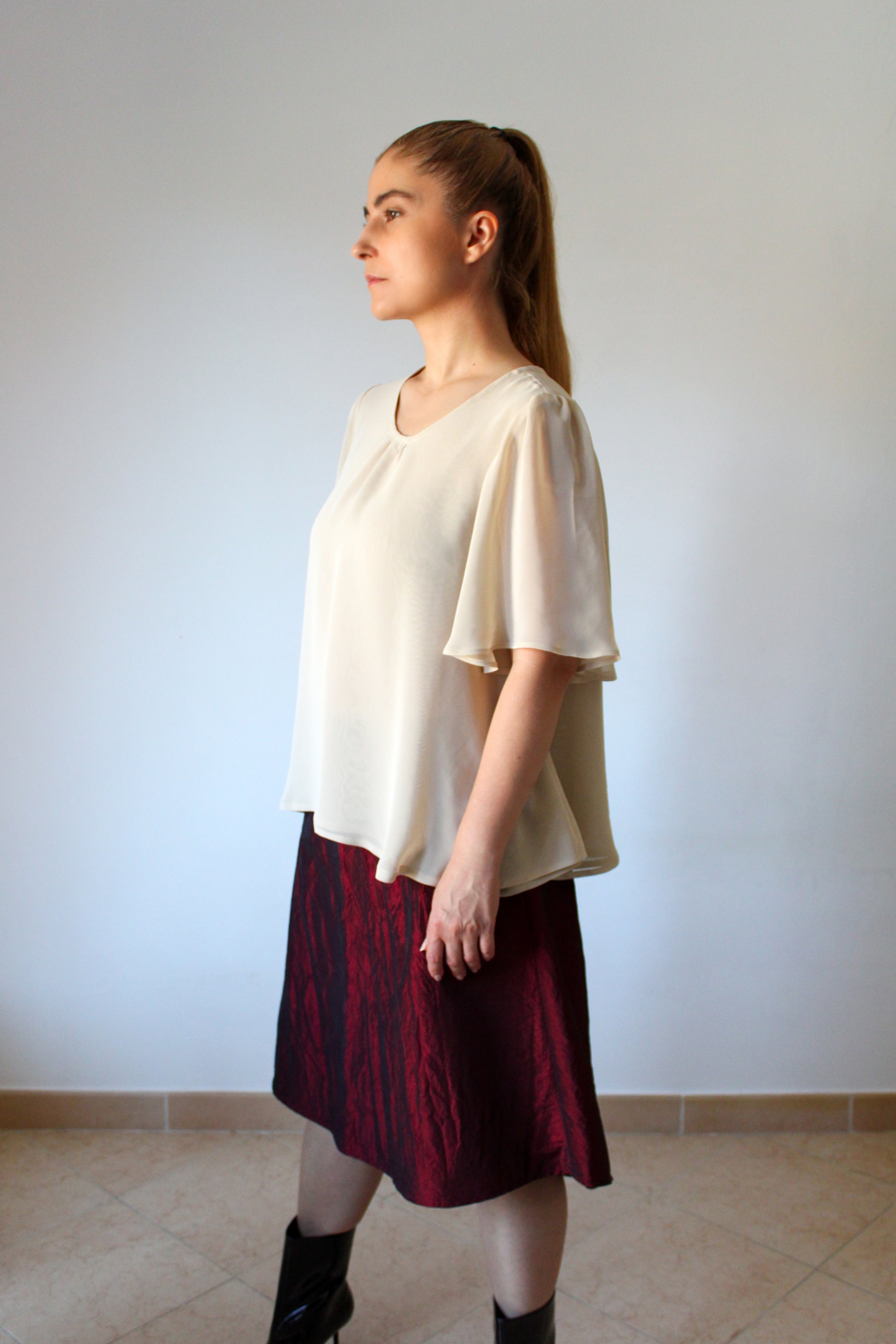 A woman photographed from the side wearing a short-sleeved chiffon blouse with a flared shape in shades of ochre and a midi Bordeaux skirt. 