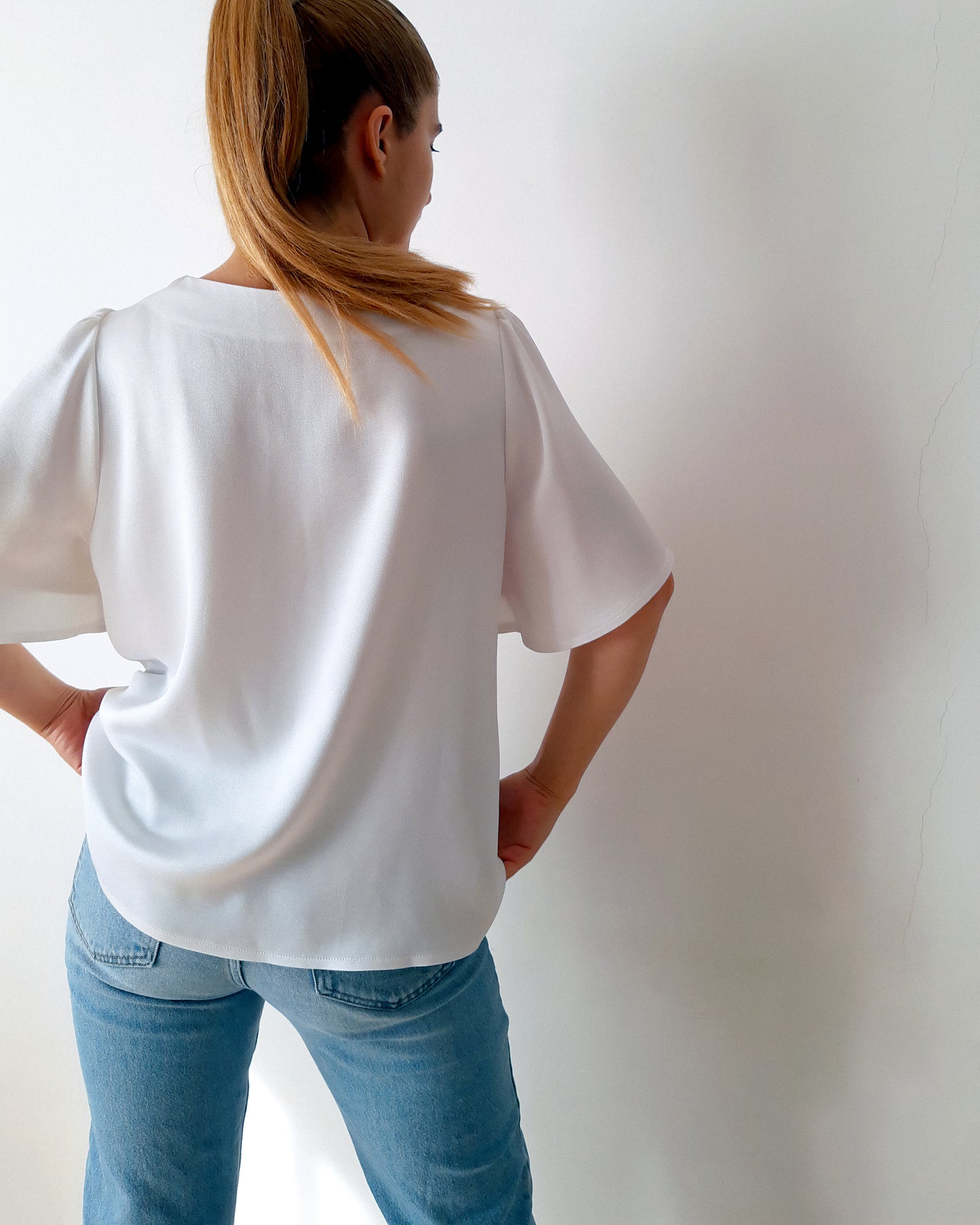 A woman is pictured from behind wearing a white crepe satin blouse with short sleeves, paired with jeans and a ponytail hairstyle.