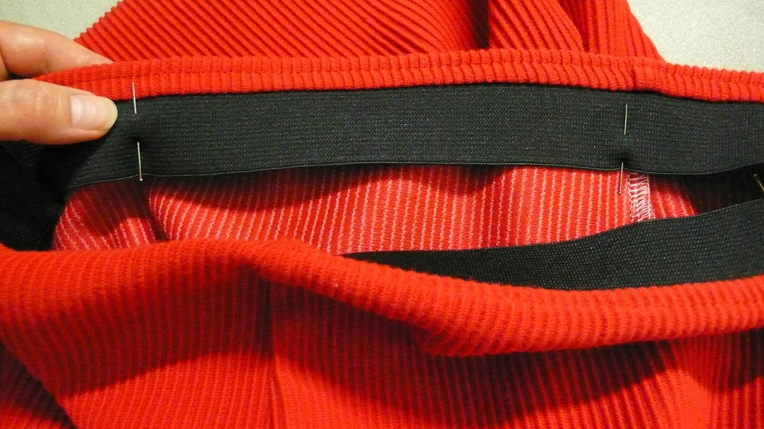 A photograph of the interior construction of the unlined skirt, specifically the waistband section. The elastic band is black, and the fabric is ribbed and red-colored.