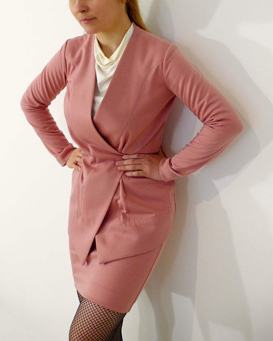 A woman is wearing an originally designed knit blazer and corresponding pencil skirt paired with a cowl neck top visible underneath. She stands against a white wall and cinches her waist with her hands.