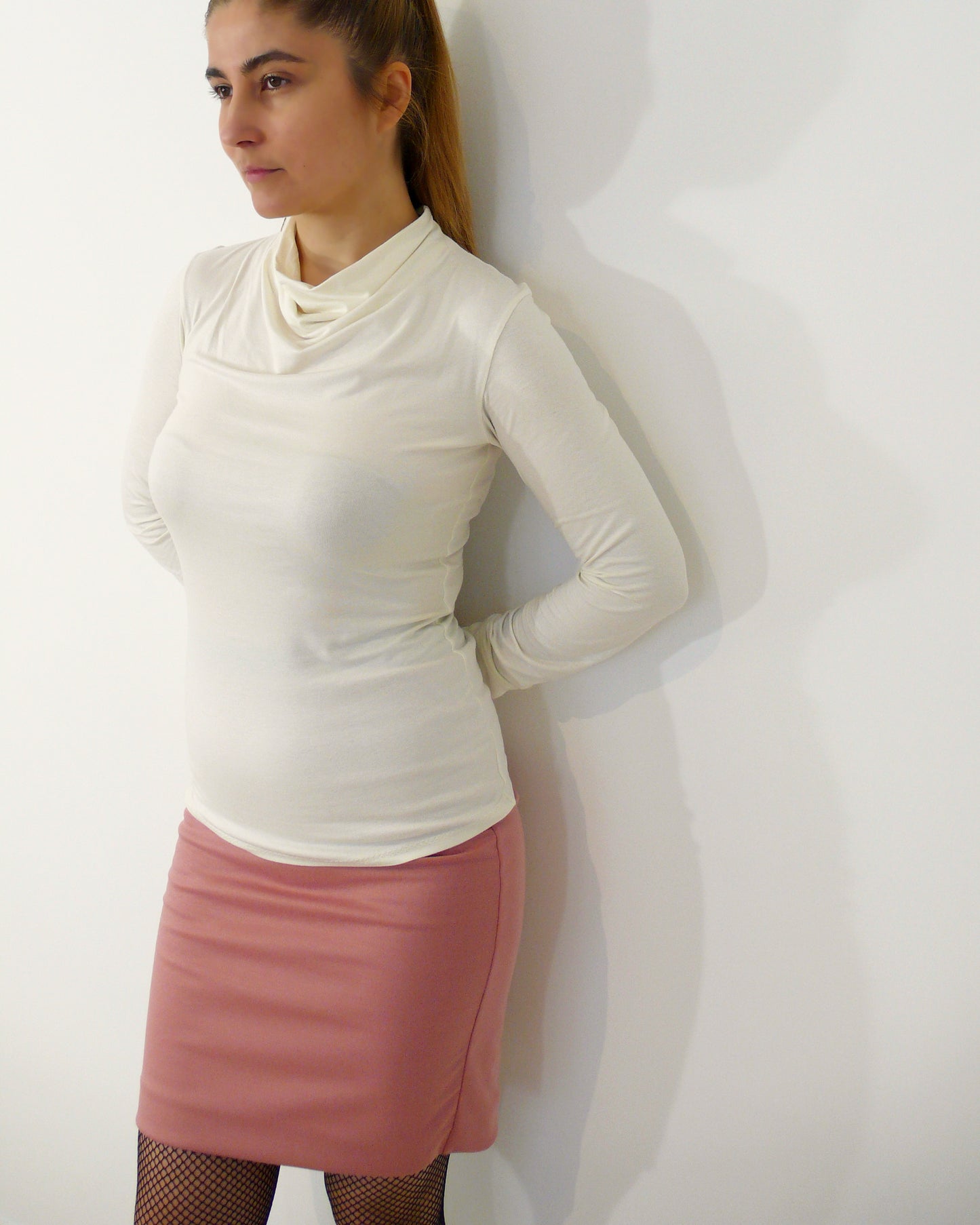A woman wearing a fitted long-sleeved cowl neck top and a bodycon skirt stands against a white wall with her arms crossed behind her back.