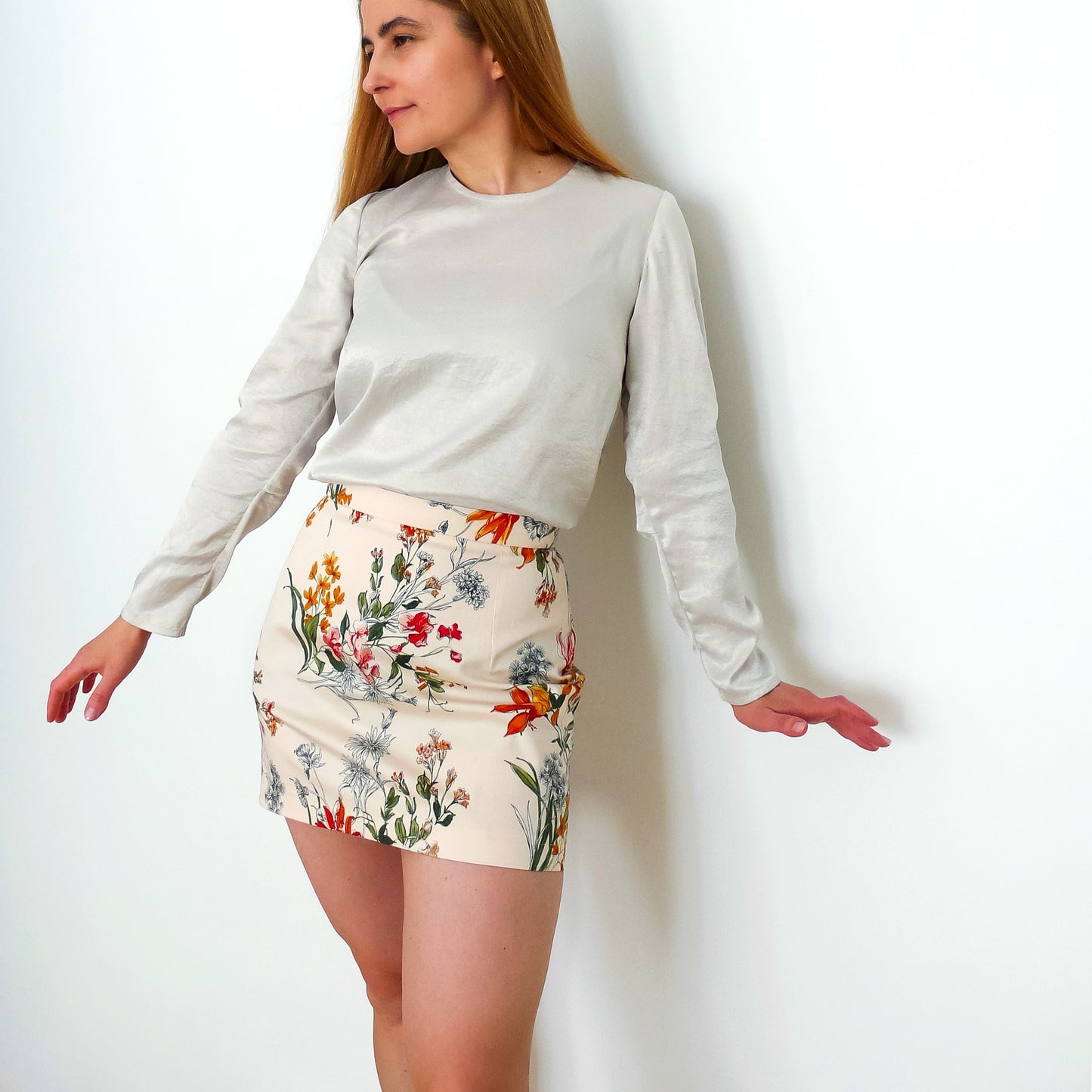 A woman posing in a light-colored satin top with long sleeves and a high-waisted floral mini-skirt. It's a front view.