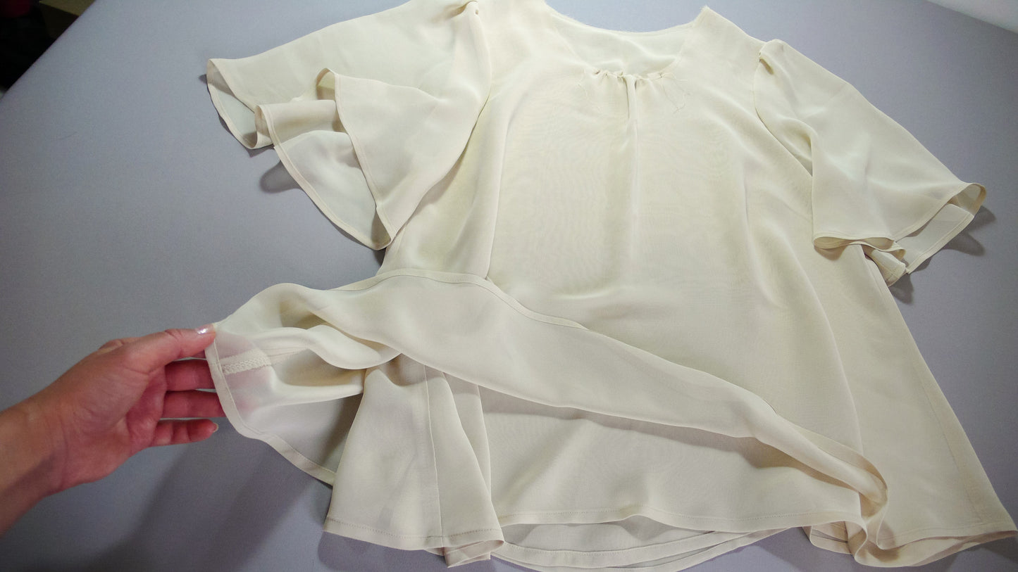 A flared blouse made of chiffon in a shade of ochre is laid out on a gray table, and a hand holds a layer of the fabric with the intent to show the seam finishing. 