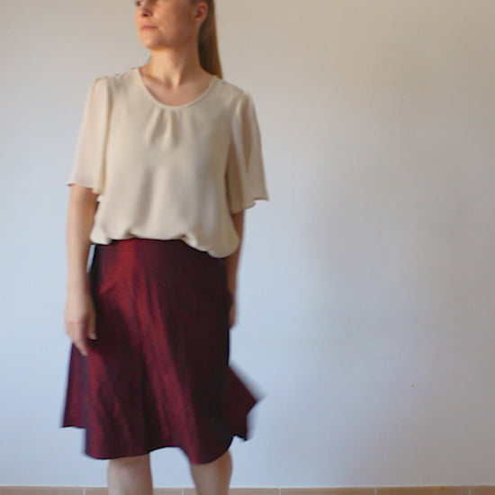 A woman models in a video a short-sleeved ochre chiffon blouse with a flared shape paired with a midi Bordeaux skirt.