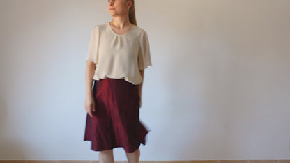 A woman models in a video a short-sleeved ochre chiffon blouse with a flared shape paired with a midi Bordeaux skirt.
